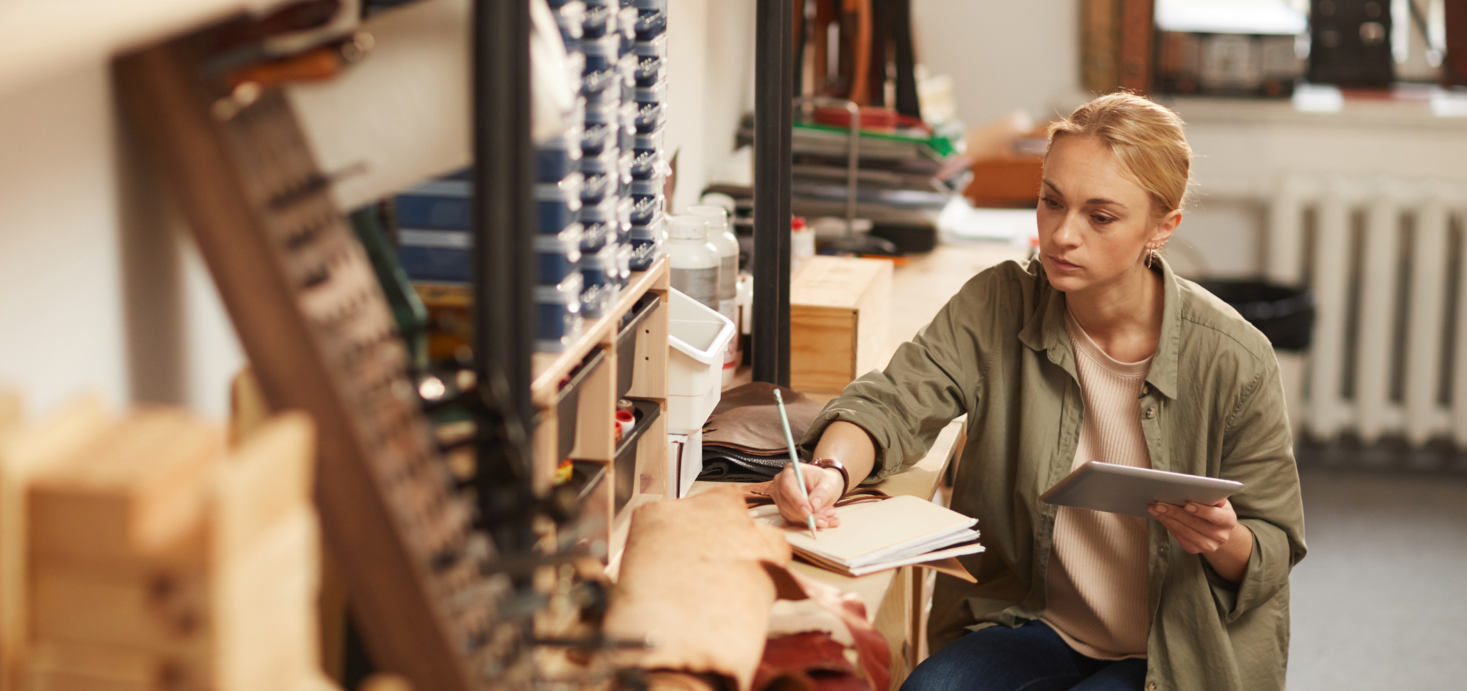 Horizontal high angle portrait of serious young woman sitting at table in craft workshop holding tablet PC and making notes in notebook